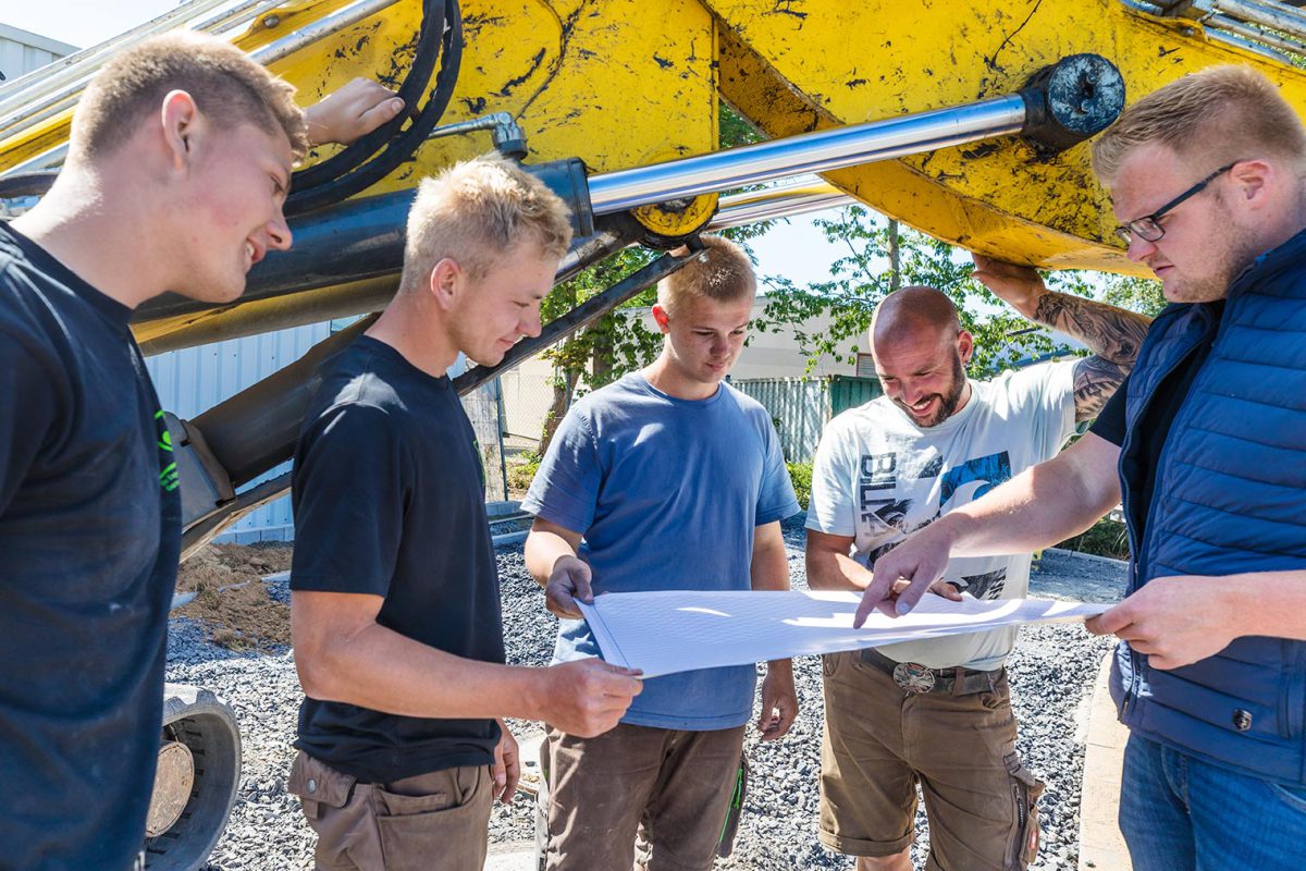 Team von Kötter Garten- und Landschaftsbau bei der Einsatzbesprechung unter einer Baggerschaufel – symbolisch für Praxisnähe und Zusammenarbeit